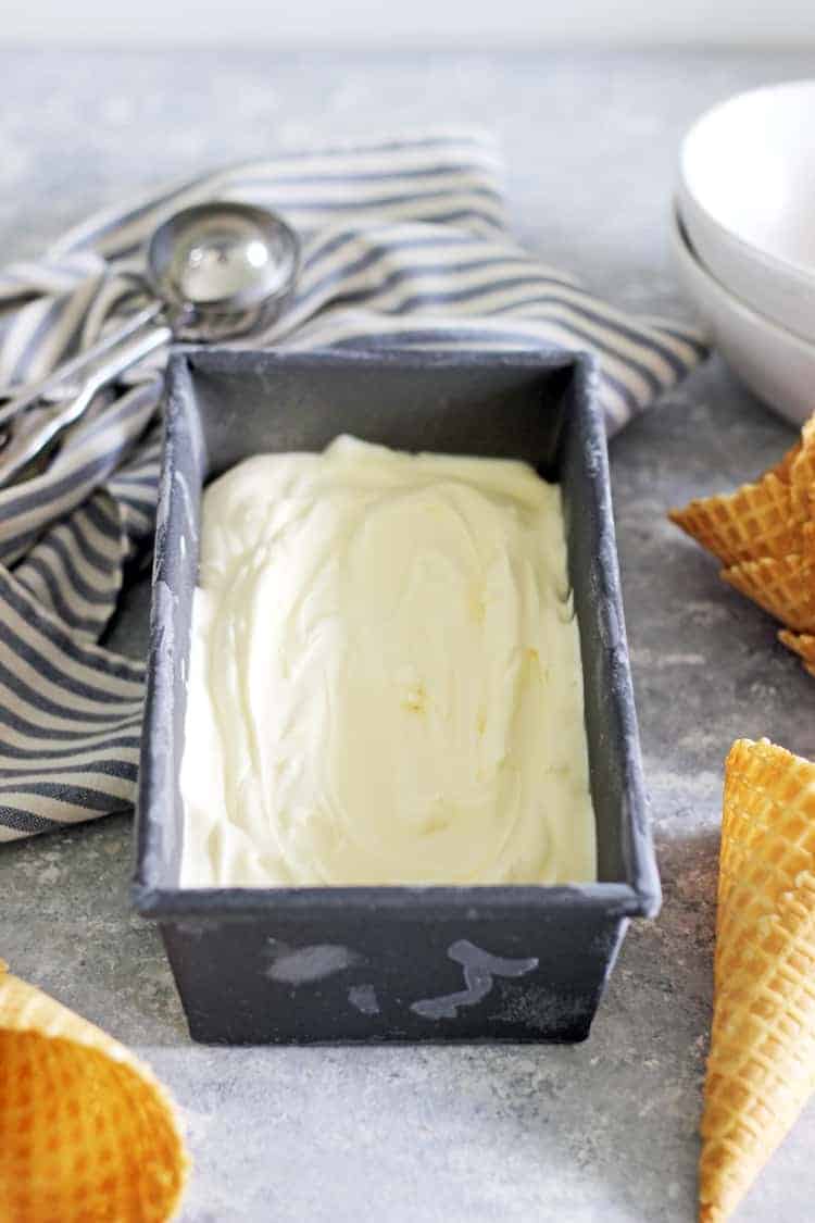 Loaf tin with vanilla ice cream in it on a grey background with waffle cones, an ice cream scoop and a tea towel in the background