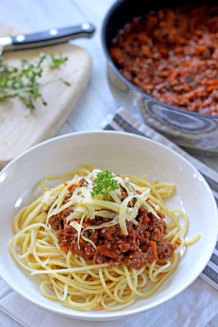 Bowl of spaghetti bolognese topped with cheese and pan of bolognese in the background