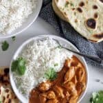 Overhead shot of white bowl with butter chicken and rice with bowl of white rice, naan bread and blue tea towel in background