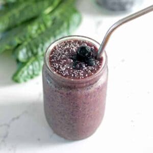 Spinach blueberry smoothie in a glass with metal straw with spinach leaves and blueberries in background cropped square