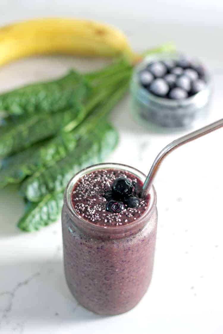 Spinach blueberry smoothie in a glass with metal straw with spinach leaves and blueberries in background