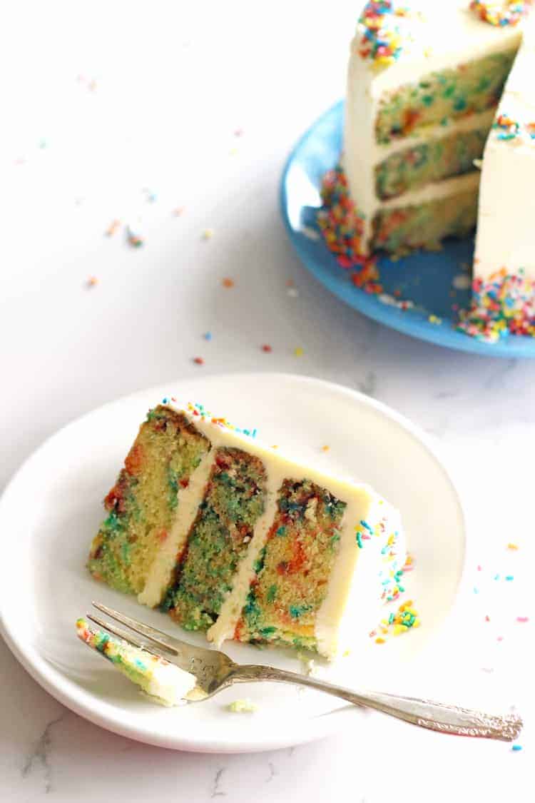 Slice of a 3 layer sprinkles layer cake on a white plate with a fork and the cake in the background