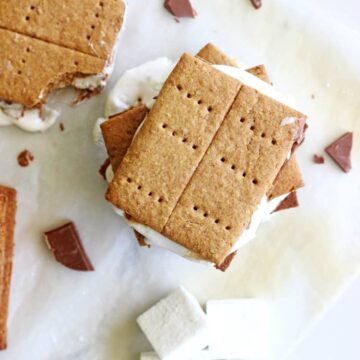 A stack of s'mores on a sheet of baking paper with marshmallows on the side and another s'more with a bite taken out