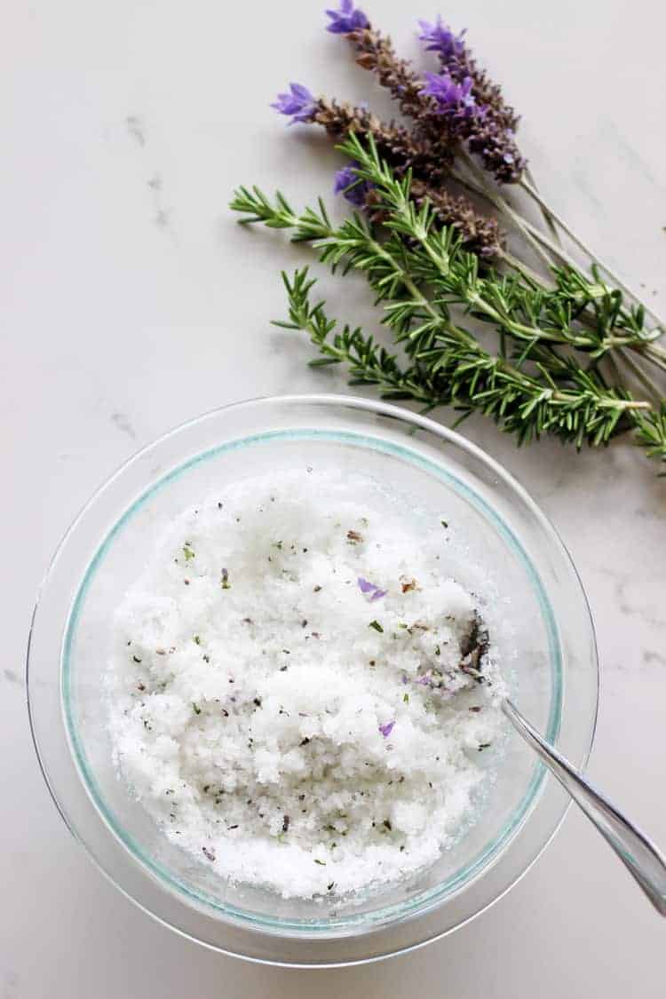 Lavender and rosemary sugar scrub in a glass bowl with lavender and rosemary on a white background