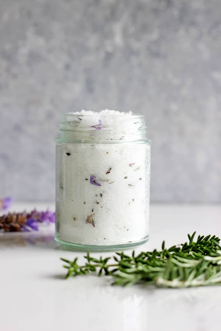 Lavender and rosemary sugar scrub in a glass jar with lavender and rosemary on a white background