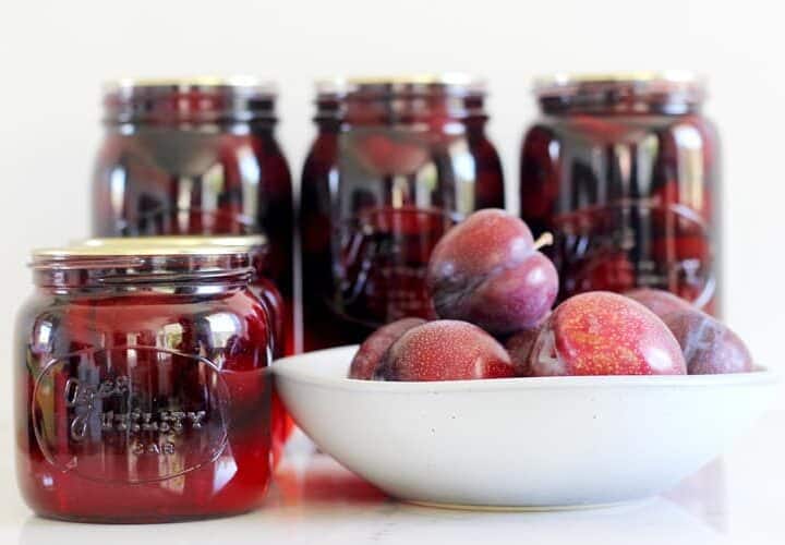 Sealed jars of bottled plums with bowl of plums in background