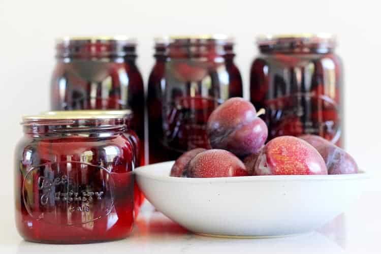 Sealed jars of bottled plums with bowl of plums in background