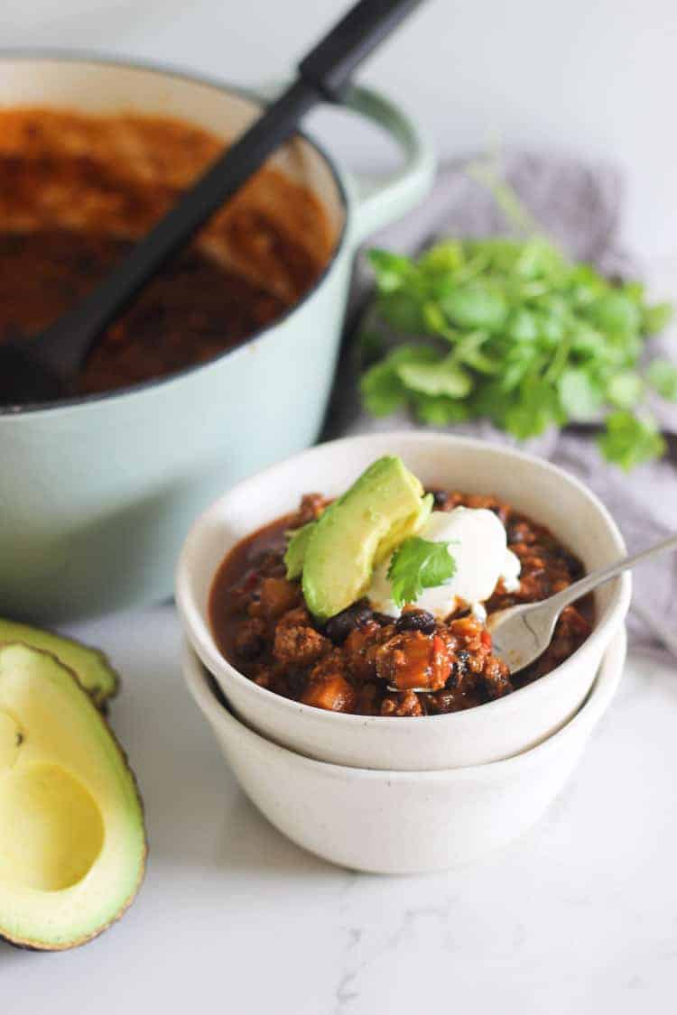 Bowl of chili with avocado, sour cream and pot in background