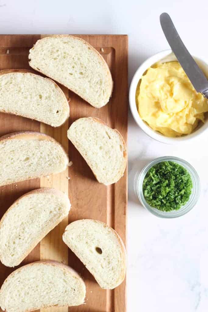 Sliced French bread on a chopping board with garlic butter and fresh parsley