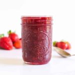 Jar of strawberry jam with strawberries in background and spoon