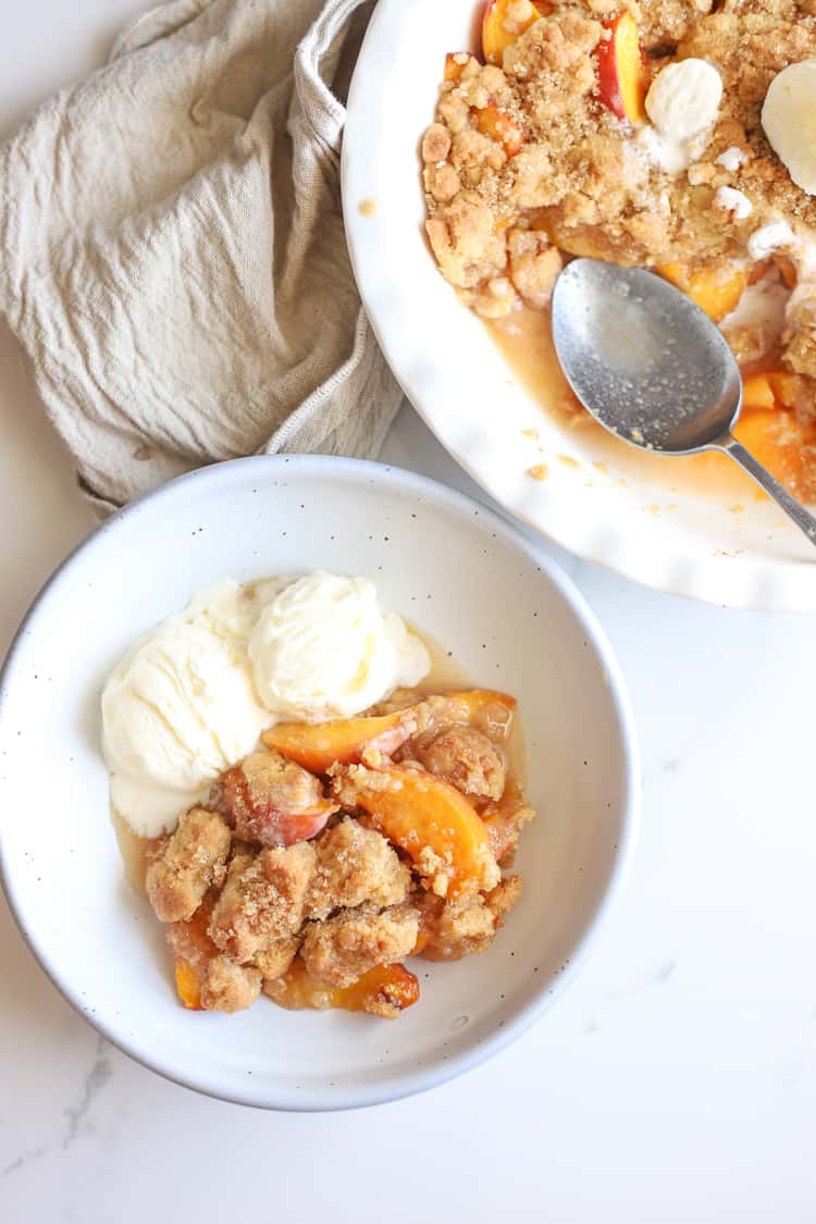 Peach cobbler with scooped vanilla icecream in a white pie dish and some in a bowl