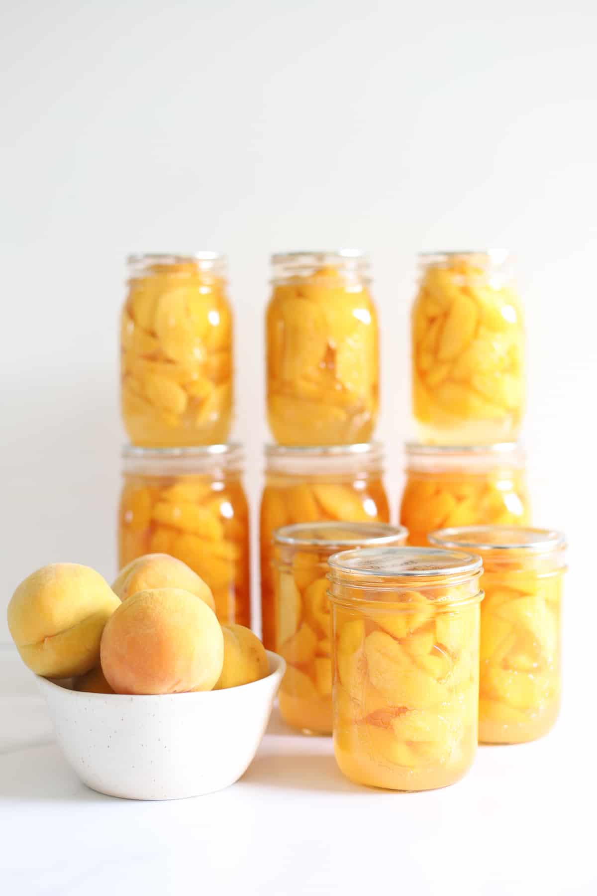 Jars of bottled peaches with a bowl of Golden Queen peaches on white background