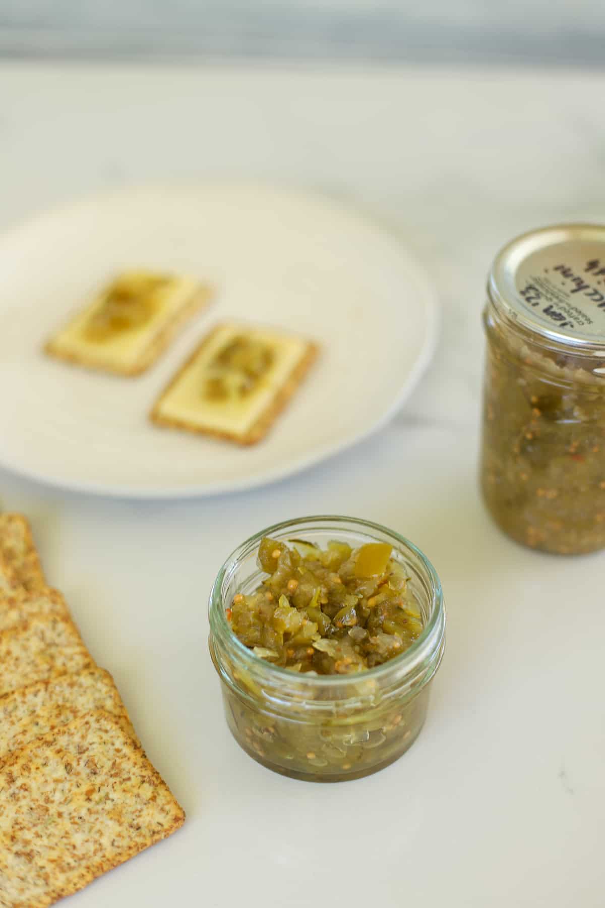 Small jar of zucchini relish with crackers on a white board